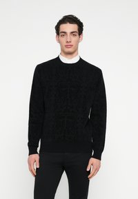 Μαύρο πλεκτό πουλόβερ με υφή λουλουδένιου σχεδίου, στρογγυλές λαιμόκοψη και ριμπ μανσέτες. Φορεμένο πάνω από ένα λευκό πουκάμισο με γιακά.