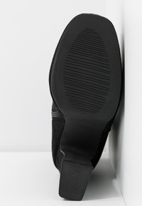 Semelle d'une botte noire à talons avec motif de texture antidérapante, posée contre une surface blanche à angle.