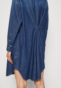 Denim overhemdjurk in donkerblauw met lange mouwen, afgeronde zoom, een rugdetail met twist dat wordt vastgezet met een knoop, en contrasterende stiksels.