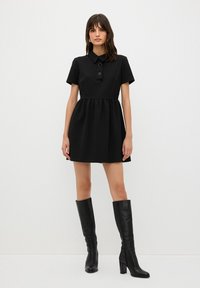 Vestido negro de manga corta con cuello tipo camisa y dos botones circulares. La falda tiene una cintura fruncida. Combinado con botas de tacón negras hasta la rodilla.