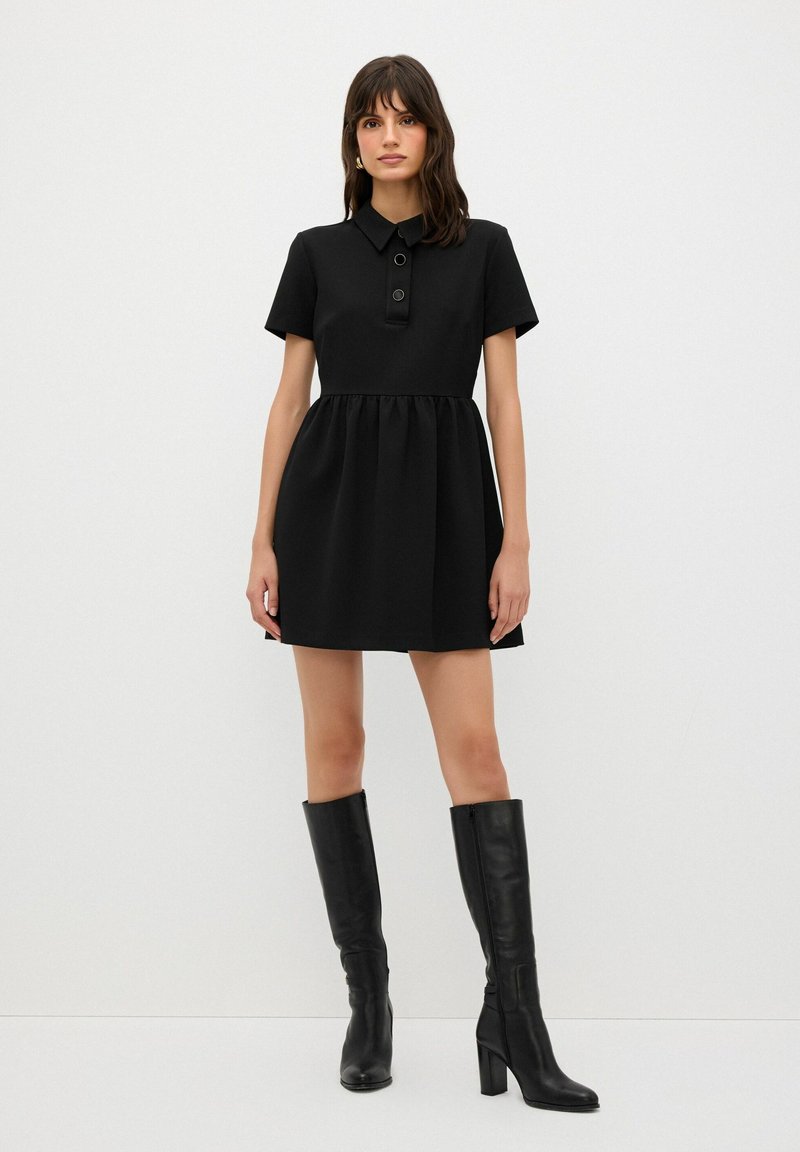 Vestido negro de manga corta con cuello tipo camisa y dos botones circulares. La falda tiene una cintura fruncida. Combinado con botas de tacón negras hasta la rodilla.