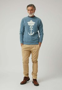 Hombre con cabello y barba canosos, vistiendo un sudadera azul con un diseño de ancla blanco, pantalones beige y botas marrones, de pie con las manos en los bolsillos.