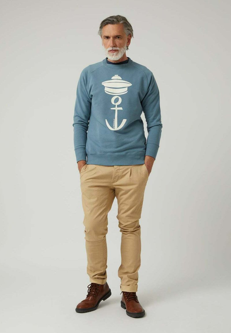 Hombre con cabello y barba canosos, vistiendo un sudadera azul con un diseño de ancla blanco, pantalones beige y botas marrones, de pie con las manos en los bolsillos.