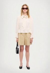 Világos bézs, hosszú ujjú blúz nyakba kötős résszel, bézs rövidnadrággal kombinálva. Fekete napszemüveg, fekete balerinacipő és egy kis fekete kézitáska.