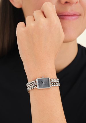 Montre bracelet en chaîne argentée avec un cadran rectangulaire gris, présentant des accents argentés et un bracelet texturé, portée au poignet.