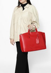 Rode totebag met een gladde, glanzende textuur; heeft dubbele handvatten en een afneembare tag. Gedragen met een oversized beige shirt en zwarte broek.