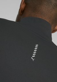 Juoda sportinė viršutinė su aukštu apykakle ir tekstūruotu audiniu. Yra balta Puma logo ir vertikalus raštas ant nugaros.