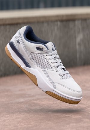 Hvid og grå atletik-sneakers med beige sål, mesh sidepanel og snørebånd, vist svævende over en betonoverflade.