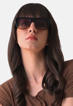 Storebrune solbriller med brune tonede linser. Rammen er kantet og tyk. Modellen har langt, bølget mørkt hår og en brun bluse.