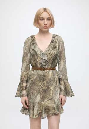 Jeune femme aux cheveux blonds courts portant une robe à manches longues à volants imprimée serpent avec une ceinture marron sur fond blanc.