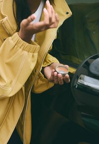 Żółta, teksturowana kurtka z elastycznymi mankietami, noszona przez osobę trzymającą małe, okrągłe metalowe pudełko obok samochodu. Widoczna gładka czarna powierzchnia samochodu.