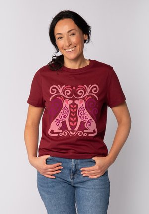 Lächelnde Frau mit dunklen Haaren trägt ein kastanienbraunes T-Shirt mit symmetrischen pinken und lila Hundemotiven und blaue Jeans, die Hände in den Taschen.