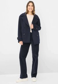 Cappotto di lana blu a doppiopetto con ampi risvolti, bottoni neri e texture morbida, abbinato a pantaloni scuri e sneakers bianche.