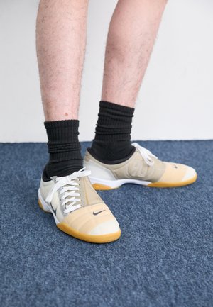 Piernas con calcetines negros y zapatos deportivos de interior Nike beige de pie sobre una alfombra azul.