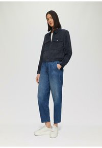 Vrouw die met één hand in haar zak staat, gekleed in een marineblauwe jas, blauwe spijkerbroek en witte sneakers, tegen een lichte achtergrond.