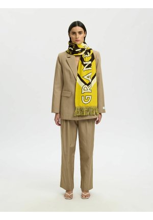 Vrouw draagt beige pak en gele sjaal met patroon, staat tegen een effen witte achtergrond, kijkt naar voren met een neutrale uitdrukking.