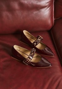 Bordeauxrote Mary Jane Schuhe aus Lackleder mit spitzen Zehen, einem Doppelriemen mit quadratischen und runden Nieten sowie niedrigen Blockabsätzen.