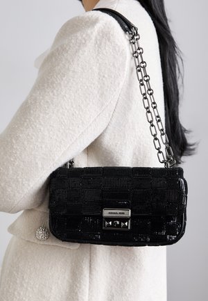 Kobieta w białym teksturowanym płaszczu nosząca czarną połyskującą torebkę na ramię Michael Kors z łańcuszkowym paskiem.