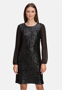 Vrouw in een knielange zwarte sequinedress met doorzichtige lange mouwen, staand tegen een effen lichte achtergrond.