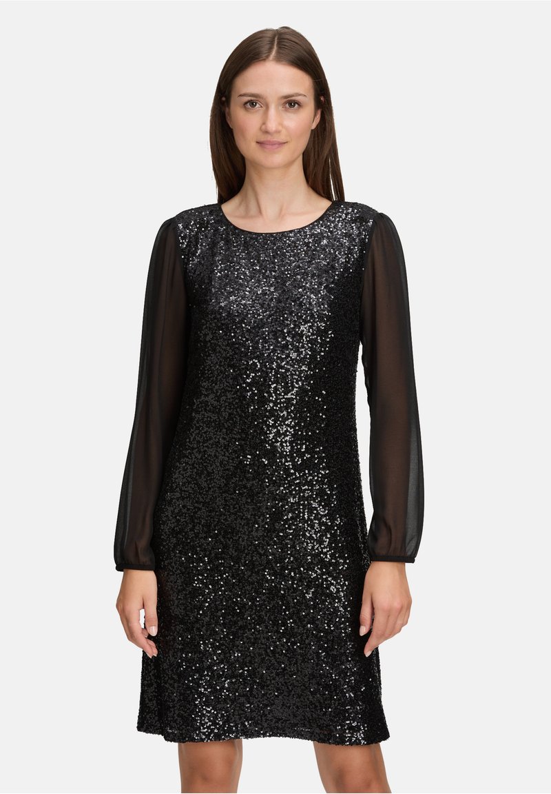 Vrouw in een knielange zwarte sequinedress met doorzichtige lange mouwen, staand tegen een effen lichte achtergrond.
