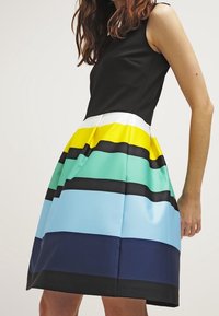 Czarna sukienka bez rękawów z dopasowanym gorsetem i rozkloszowaną spódnicą, ozdobiona poziomymi paskami w kolorze żółtym, zielonym, niebieskim i granatowym. Gładki materiał.