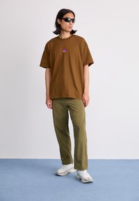 Brun bomull T-skjorte med korte ermer, med en liten lilla logo. Kombinert med olivengrønne bukser og hvite sneakers. Enkel bakgrunn.