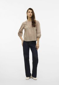 Top marrone chiaro e testurizzato con maniche a tre quarti e scollatura arrotondata, abbinato a jeans a gamba larga blu scuro. Calzature lisce completano il look.