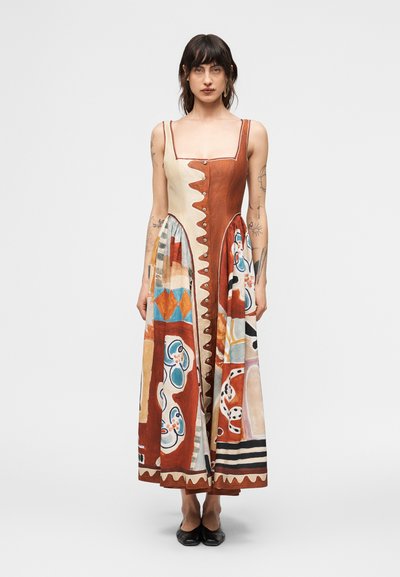Femme portant une robe maxi sans manches avec des motifs abstraits multicolores marron et crème, debout devant un fond clair uni.