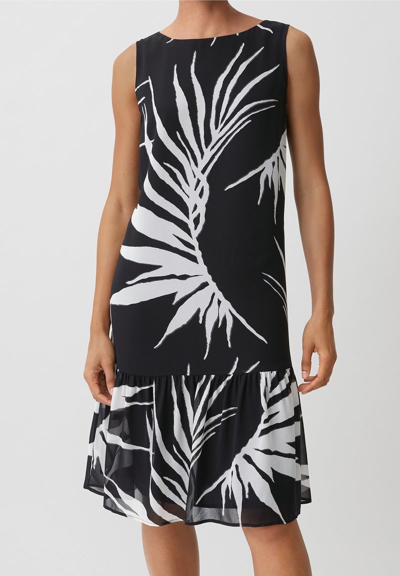 Robe noire sans manches avec grand imprimé de feuilles tropicales blanches et ourlet à volants, portée par une personne avec les bras détendus le long du corps.