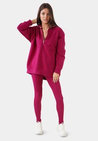 Sudadera magenta con cremallera y corte relajado, combinada con leggings a juego. Presenta una tela suave y puños acanalados. Llevada con zapatillas blancas.