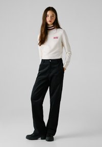 Weißer Pullover mit rot- und kastanienbraun gestreiftem Kragen, mit dem Logo "Red Bull Racing". Schwarze weite Hose und schwarze Schuhe runden das Outfit ab.