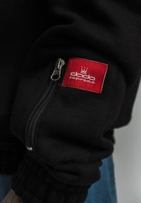 Schwarzer Sweatshirt mit einer Reißverschlusstasche am Ärmel. Mit einem roten Etikett mit weißem Text und einem Kronenlogo. Weicher Stoff und gerippte Bündchen.