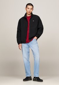 Zwarte bomberjack met een hoge kraag, rode shirt eronder, lichtblauwe spijkerbroek en zwarte sneakers. Opvallende details van rits en manchetten.