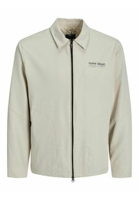 Giacca beige chiaro con chiusura a zip, colletto aperto e tasche laterali. Presenta un logo "core dept." in grigio scuro sul davanti.