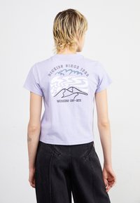 Maglietta corta lavanda con grafica sul retro di montagne e nuvole, con il testo "DICKIES SINCE 1922" e "WORKING OFF-SITE." Maniche corte.