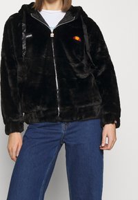 Donna che indossa un hoodie con zip in pelliccia sintetica nera e loghi, una maglietta bianca e jeans blu, in piedi contro uno sfondo chiaro e neutro.