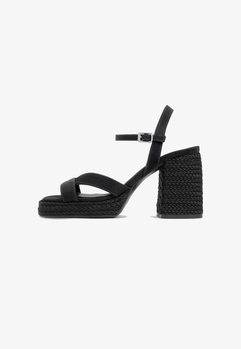 Sandale noire à talon haut avec plateforme tressée et talon bloc, bride de cheville avec boucle argentée, et brides croisées à l’avant.