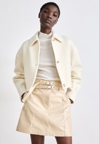 Veste cropped blanche texturée avec de gros boutons associée à une jupe en cuir beige taille haute avec un design à double ceinture.