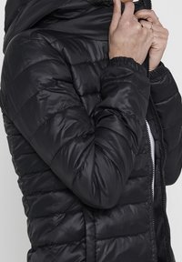 Black puffy jacket with a hood, quilted texture, full front zipper, and gathered cuffs. Features a smooth, shiny fabric finish.