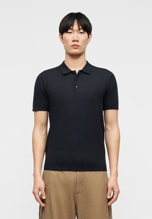 Jeune homme aux cheveux noirs portant un polo noir à manches courtes et un pantalon beige, faisant face à l'avant avec une expression neutre.