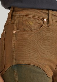 Jean en denim marron avec des rivets en cuivre et un petit logo brodé au-dessus de la poche avant, porté avec un haut foncé.