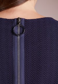 Mörkblått texturerat tyg med ett fiskbensmönster, med en synlig silverfärgad dragkedja med ett runt draghandtag vid baksidan av halsringningen.