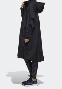 Černá oversized deštník s kapucí, vyrobená z texturované tkaniny a zdobená knoflíkovými detaily. V kombinaci se pruhovanými legínami a černými sportovními botami.