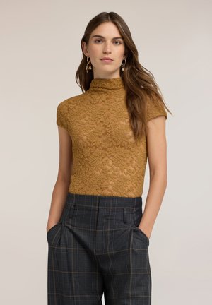 Mujer con cabello largo y castaño que lleva pendientes dorados colgantes, una blusa de encaje de manga corta color mostaza y pantalones oscuros de cuadros de talle alto con las manos en los bolsillos.