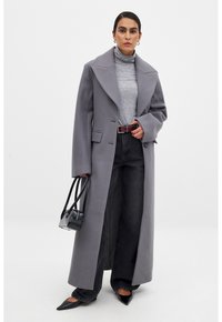 Langer grauer Wollmantel mit übergroßen Revers, getragen über einem hellgrauen Rollkragenpullover. Kombiniert mit dunklen, weit geschnittenen Jeans und spitzen schwarzen Schuhen. schwarze Handtasche.