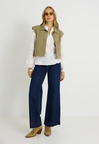 Chaleco corto verde oliva sobre una blusa blanca bordada, combinado con vaqueros anchos de pierna ancha en azul oscuro y botines de tacón en color beige.