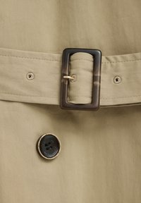 Close-up of beige fabric belt with dark rectangular buckle and a black button stitched with "Polo Club" text.