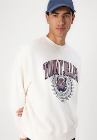 Witte sweatshirt met "TOMMY JEANS" tekst en tijgerafbeelding. Voorzien van lange mouwen, geribbelde kraag en zachte textuur. Draagt een blauwe pet.