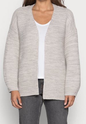 Femme portant un cardigan en maille gris clair par-dessus un t-shirt blanc et un jean gris foncé, debout devant un fond uni.