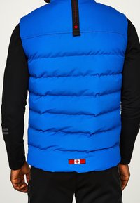 Blaues gesteppte Weste, aus synthetischem Material, horizontale Nähte, hoher Kragen, schwarze Akzente, mit einem Kanada-Flaggen-Patch am Saum.